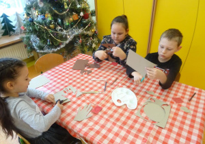 Troje dzieci siedzi przy stole, obrysowują swoje dłonie na kartce i wycinają szablony.